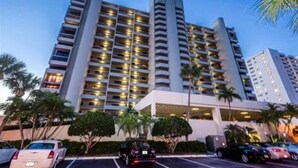 A multi-story hotel with a modern design, featuring balconies on each floor, a parking area with several cars, and palm trees in the foreground.