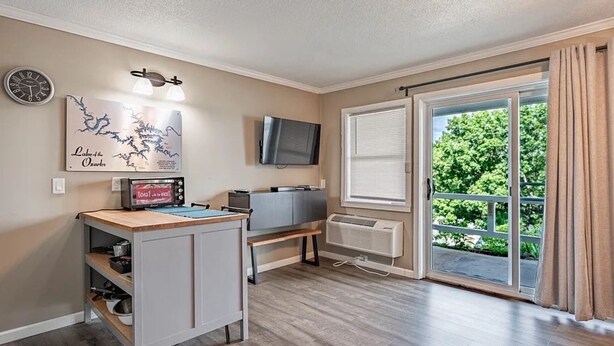 A modern living room with a flat-screen TV, a dining area with a microwave, and a large window with a view of greenery.