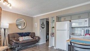 A modern living room with a brown leather sofa, a round mirror, and a kitchen area with white cabinets and a microwave.