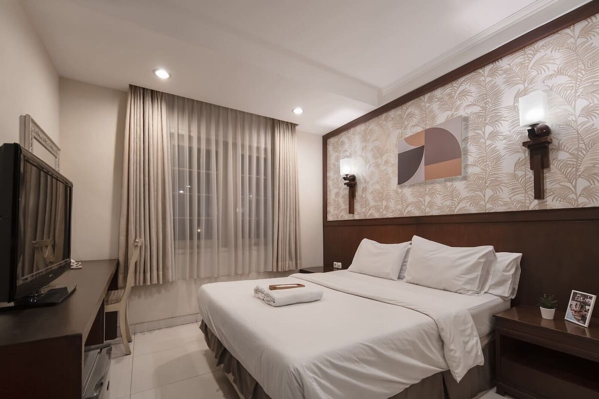 A hotel room with a bed, a TV, a desk, and a window with curtains.