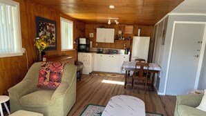 A cozy room with a sofa, ottoman, and a small table with a rug. A kitchen area with cabinets and a refrigerator is visible in the background. - Cozy, comfortable and close to CBD (Whakatane)