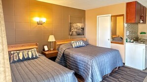 A hotel room with two beds, a nightstand, a lamp, a painting, a mirror, and a kitchenette. - Athabasca Hillside Motel (Athabasca)