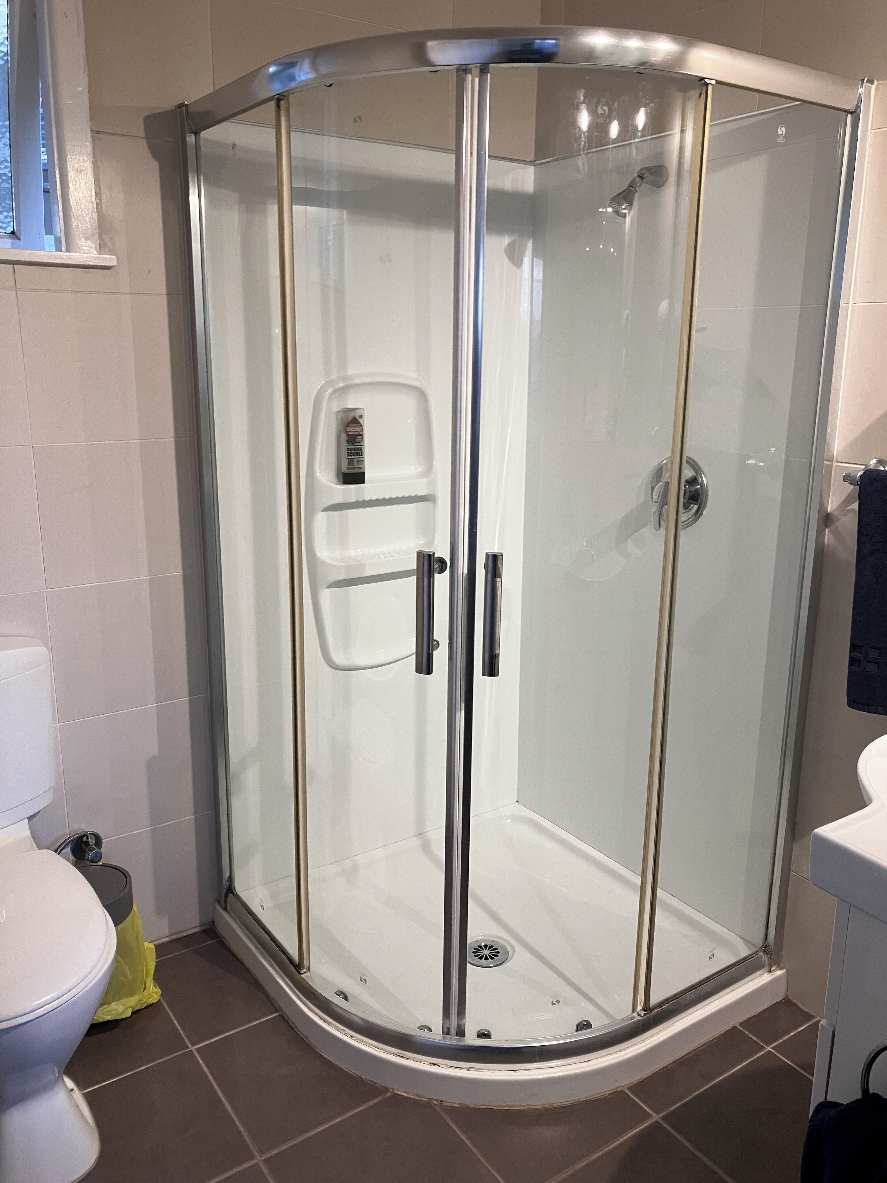 A glass-enclosed shower with a white tiled floor, a toilet, and a sink with a towel hanging on the wall.
