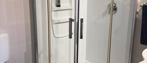 A glass-enclosed shower with a white tiled floor, a toilet, and a sink with a towel hanging on the wall.