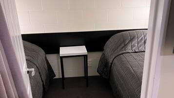 Quarto pequeno e estreito com duas camas de solteiro, uma mesinha e uma parede de tijolos brancos.