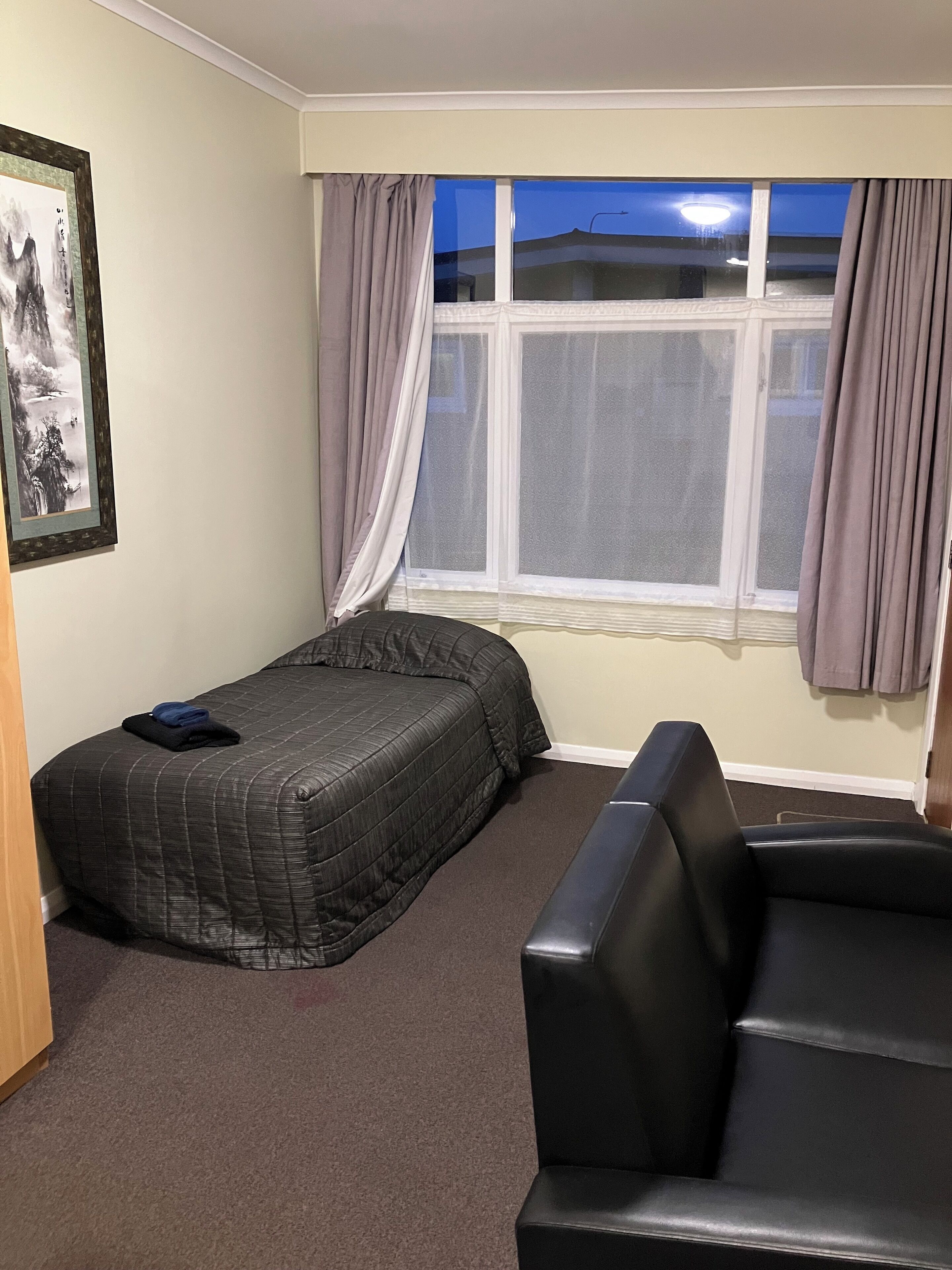A room with a bed, a chair, a window with curtains, and a framed picture on the wall.