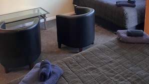 Une chambre d’hôtel moderne dotée d’un téléviseur à écran plat fixé sur une étagère en verre, d’un lit confortable avec une couette de couleur foncée, d’un petit pouf rond et d’un coin salon accueillant avec un fauteuil rembourré.
