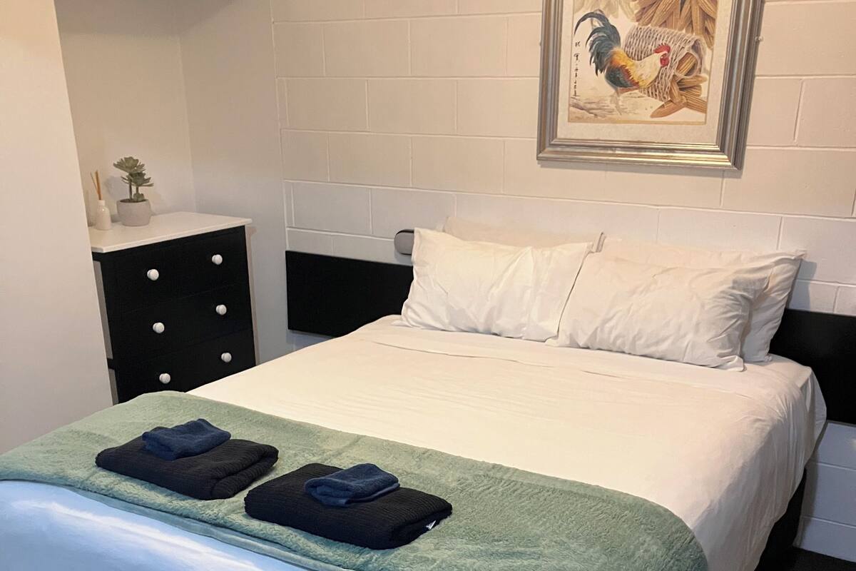A neatly made bed with a green blanket and two folded towels, a black nightstand with a drawer, and a framed picture on the wall.