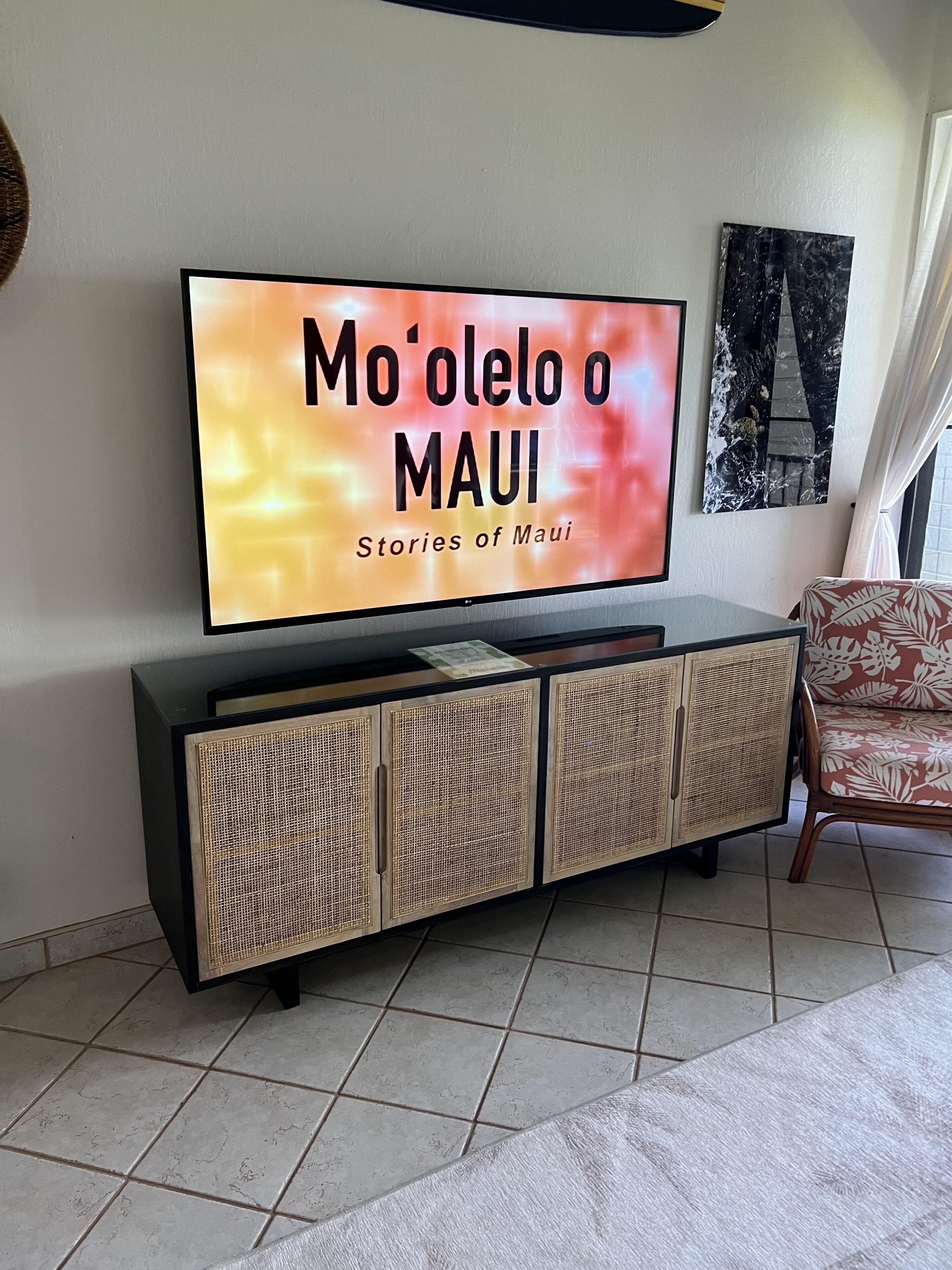 Een flatscreen televisie op een zwarte tv-standaard met een gevlochten kastje, waarop staat 'Mo'olelo o MAUI - Stories of Maui'.