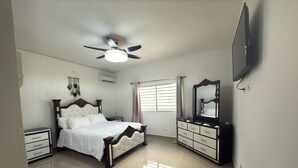 Une chambre à coucher comprenant un lit, une commode, un téléviseur, un ventilateur de plafond et une fenêtre avec des rideaux.