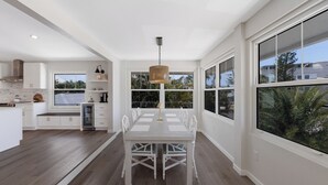 A modern kitchen with white cabinetry, a dining table with chairs, and large windows offering views of the outdoors. - Luxury Beach Home•Steps to Beach•Excellent Location•Sunsets (Holmes Beach)