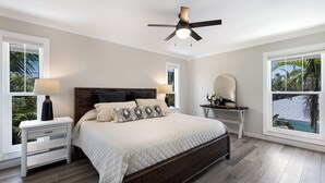 A bedroom with a bed, nightstands, a desk, a ceiling fan, and large windows with views of palm trees. - Luxury Beach Home•Steps to Beach•Excellent Location•Sunsets (Holmes Beach)