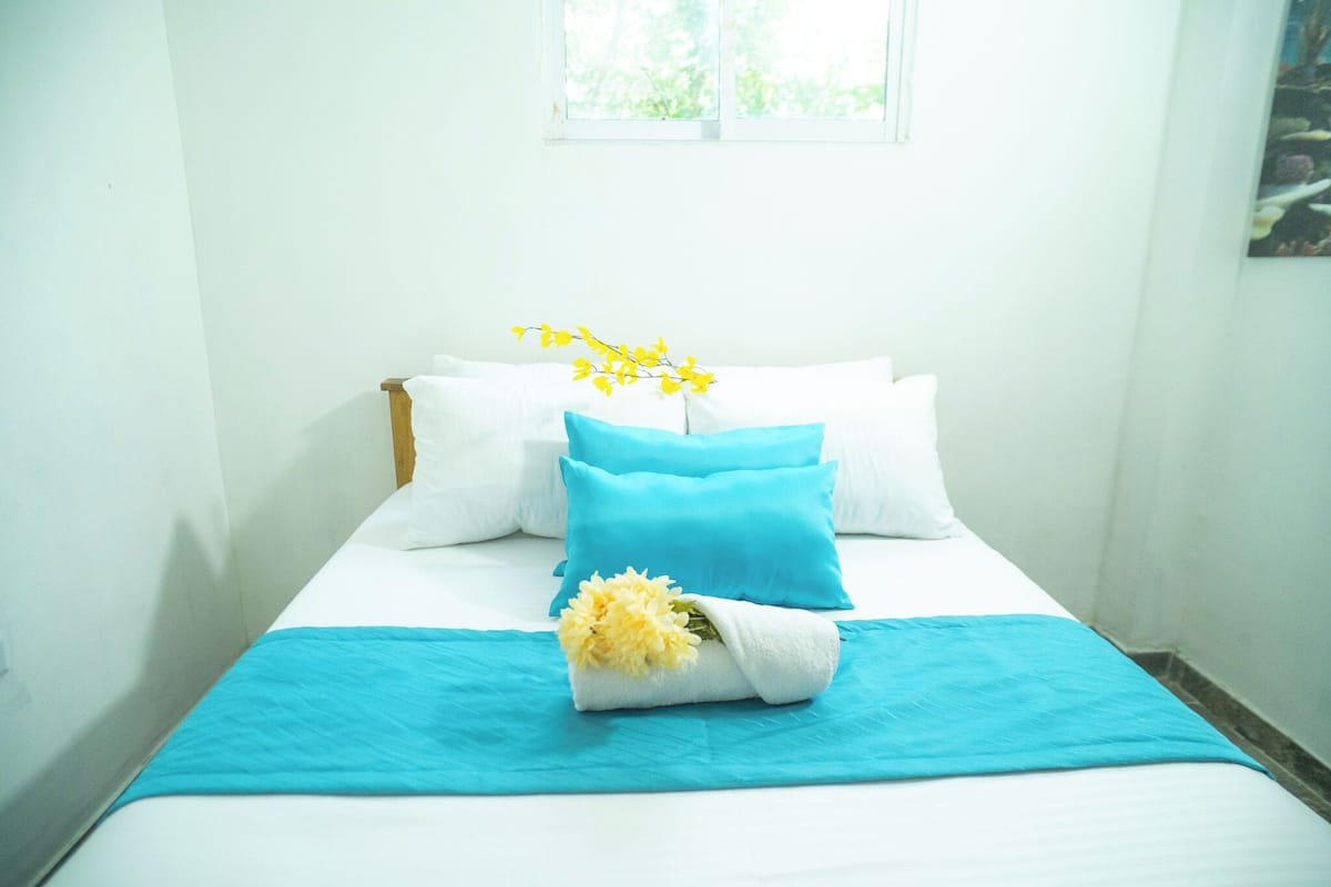 Ein ordentlich bezogenes Bett mit weißer und blauer Bettwäsche, einer blauen Decke und einem Strauß gelber Blumen.