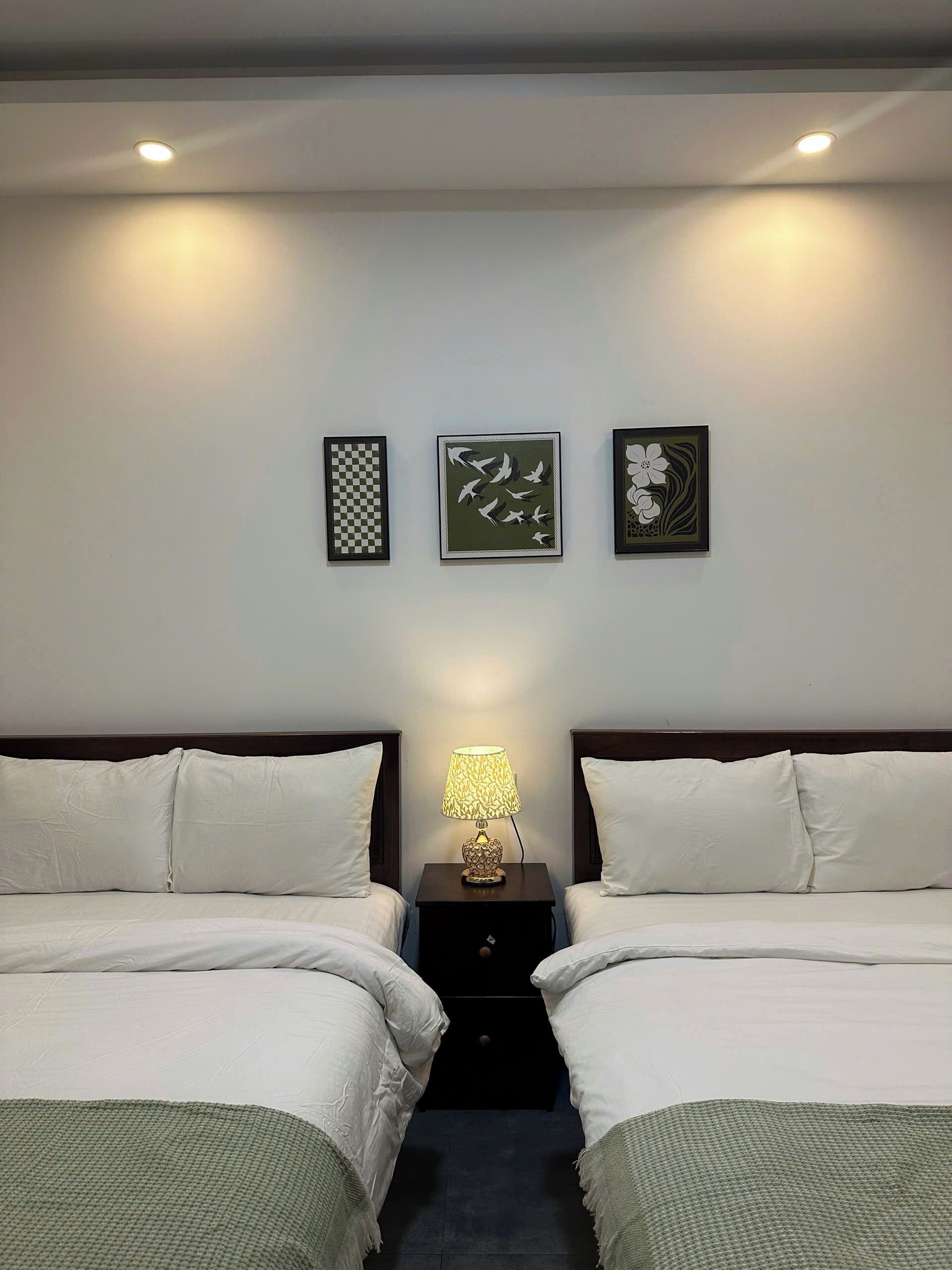 A hotel room with two beds, a nightstand with a lamp, and three framed artworks on the wall.