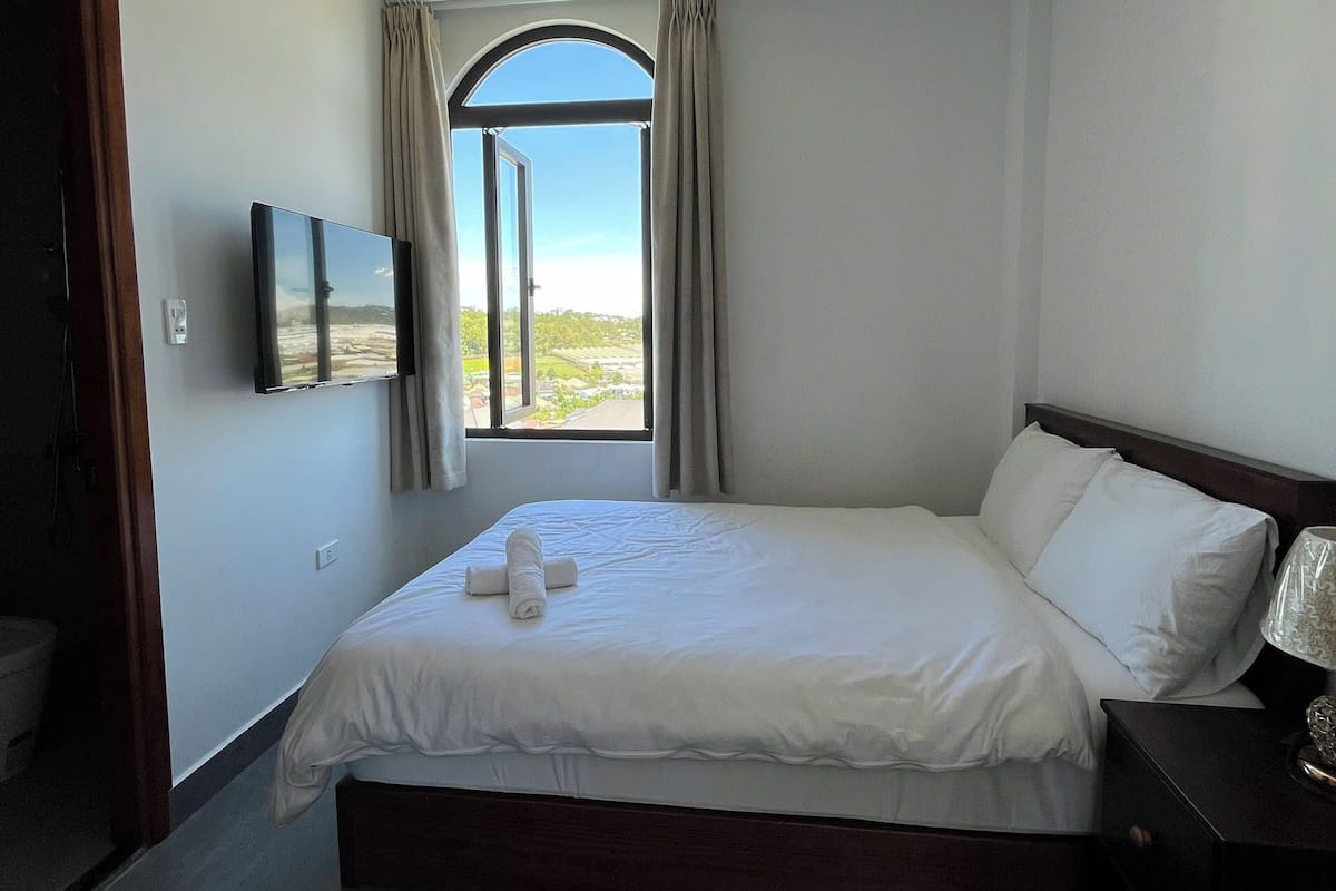 A bedroom with a large bed, a TV mounted on the wall, and a window with curtains.