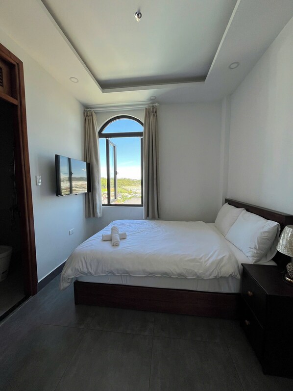 A bedroom with a large bed, a TV mounted on the wall, and a window with curtains.