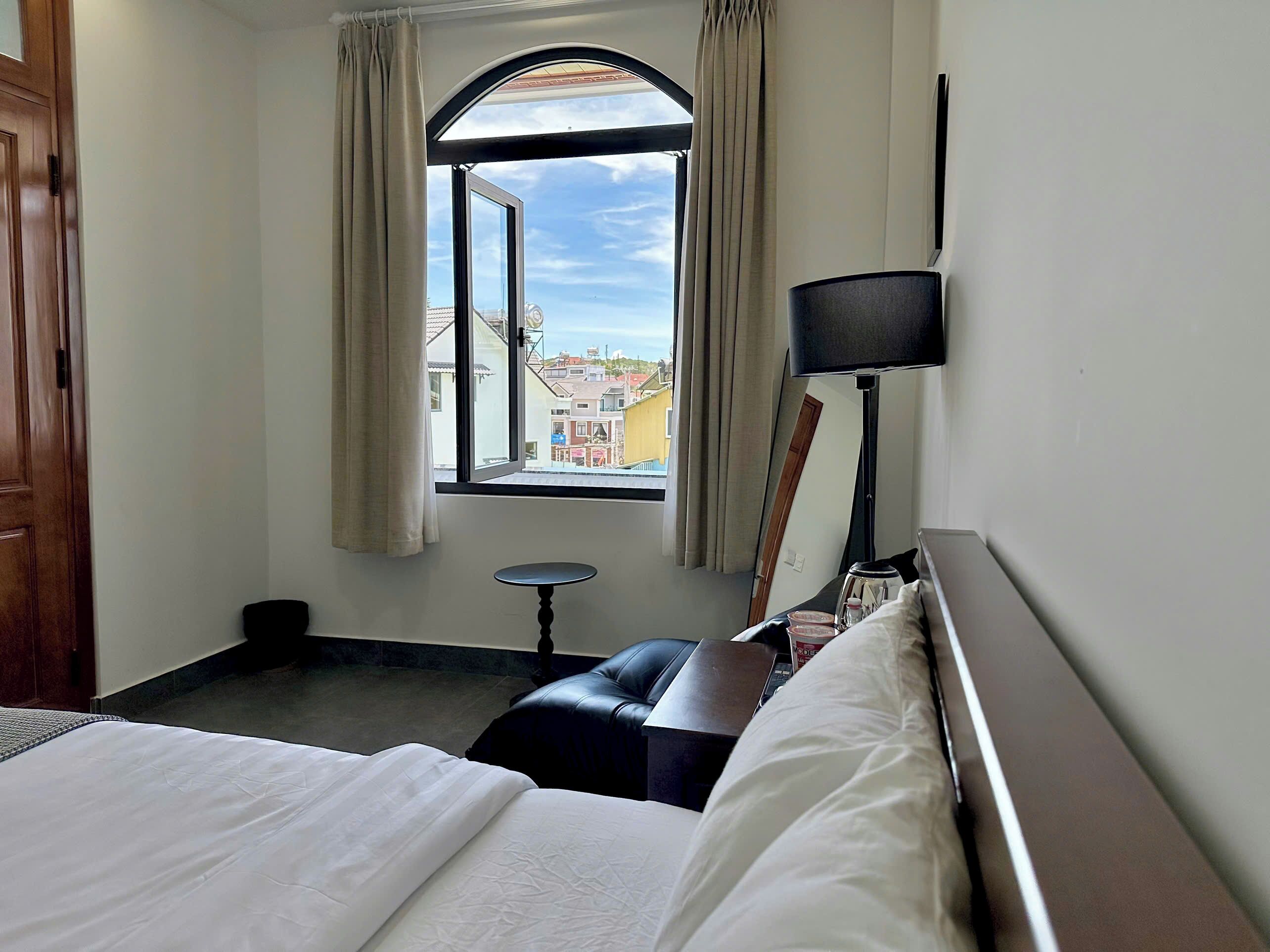 A hotel room with a bed, a small table, a chair, a lamp, and a window with curtains.