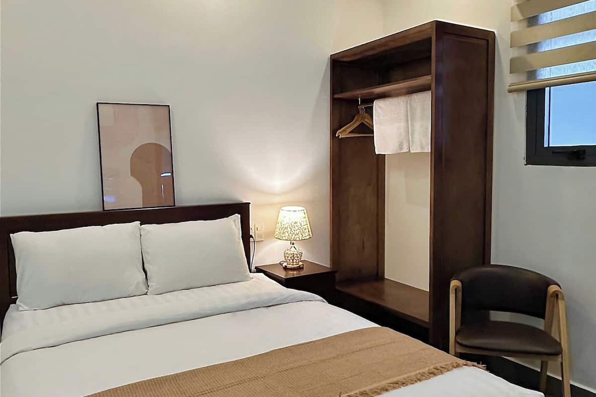 A hotel room with a large bed, a wooden wardrobe, a chair, a bedside table with a lamp, and a framed picture on the wall.