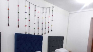 Une chambre d’hôtel avec trois lits, chacun doté de draps blancs et d’une tête de lit bleue. Une guirlande de pompons colorés est accrochée au mur.