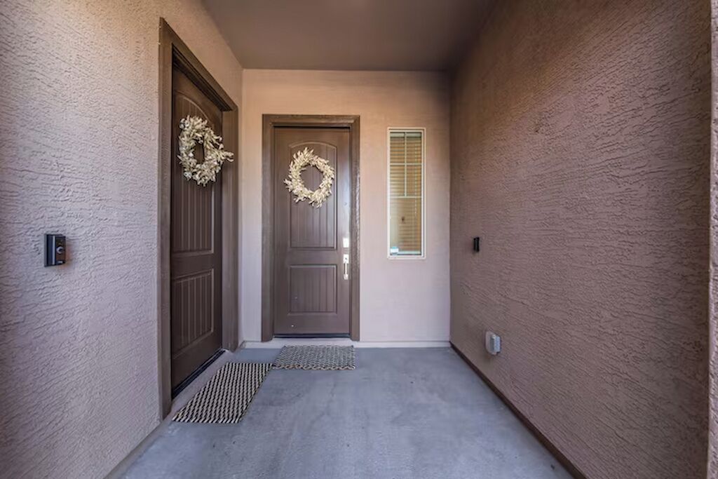 A hallway with two doors, one with a wreath, a window with blinds, and a doormat.