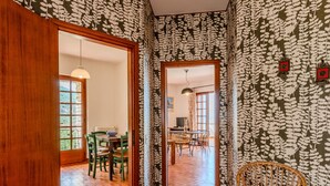 A room with patterned wallpaper, a wooden floor, a dining area with chairs and a table, and a wicker chair. - Papandrea by Interhome (Brenzone)