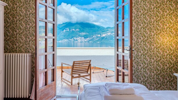 A bedroom with a wooden bed, a chair, and a view of a lake and mountains through open doors. - Papandrea by Interhome (Brenzone)