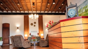 A wooden reception desk with a clock, two armchairs, a chandelier, and a stone wall with a painting. - The Golden Legacy Hotel (Nuwara Eliya)