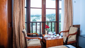Una habitación con una mesa de madera dispuesta para el té, dos sillas, un jarrón con flores y vistas a edificios a través de amplias ventanas.