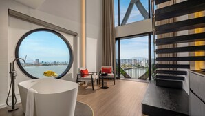 A modern bathroom with a bathtub, a round window, and a view of a cityscape. - Caro Premium Danang Hotel (Da Nang)