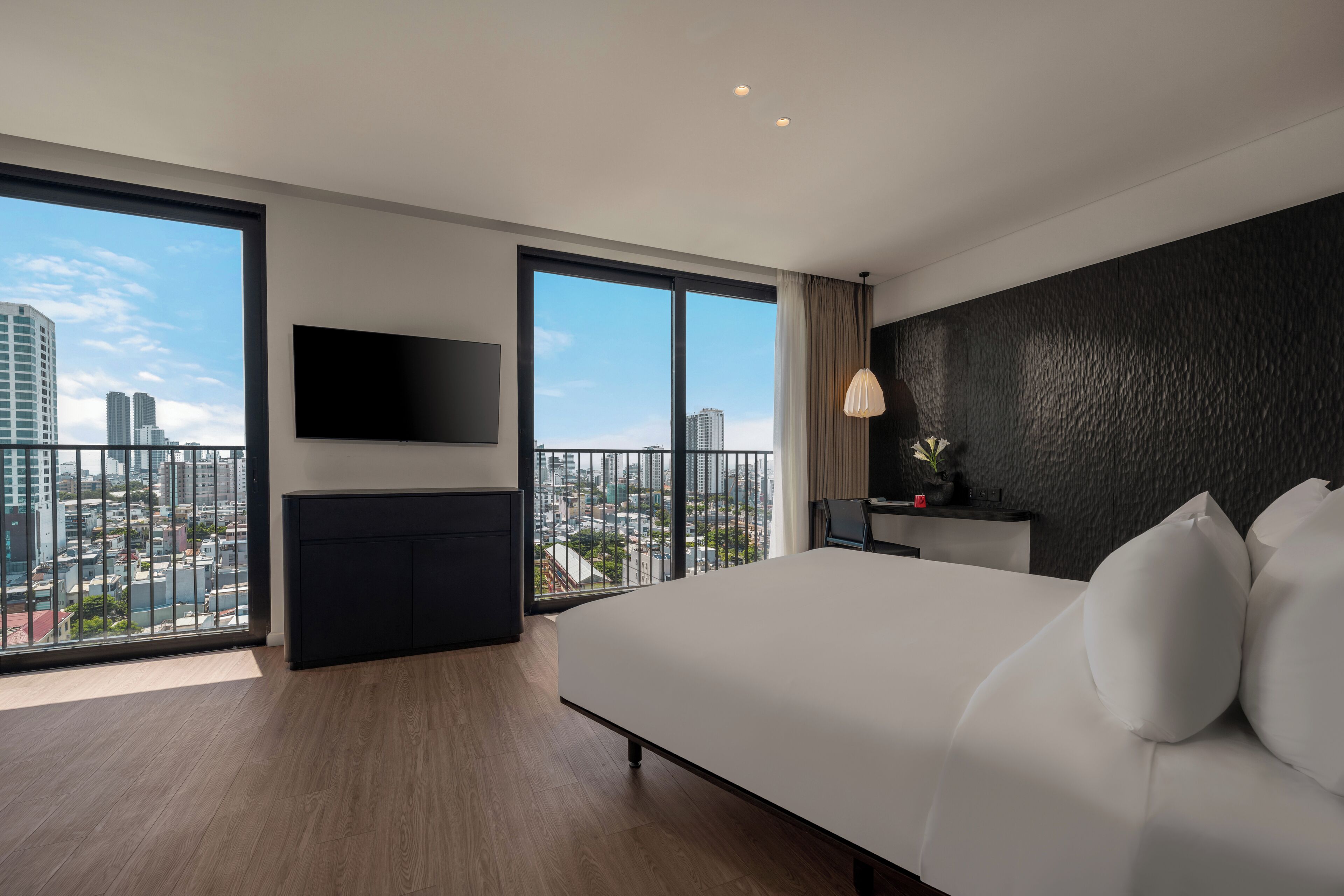 Un hotel moderno con una cama grande, un televisor de pantalla plana en la pared, una mesita de noche pequeña y vistas de la ciudad a través de amplias ventanas.