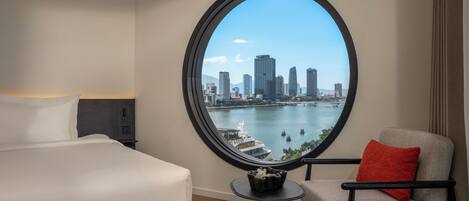A hotel room with a large round window offering a city skyline view, a bed, a chair, and a small table with a decorative item.