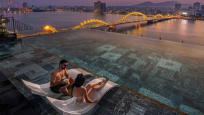 Een koppel ontspant op een drijvend platform in een zwembad, met een stadsbeeld en een brug op de achtergrond.