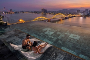 Ein Paar entspannt auf einer schwimmenden Plattform in einem Pool, im Hintergrund eine Stadtlandschaft und eine Brücke.