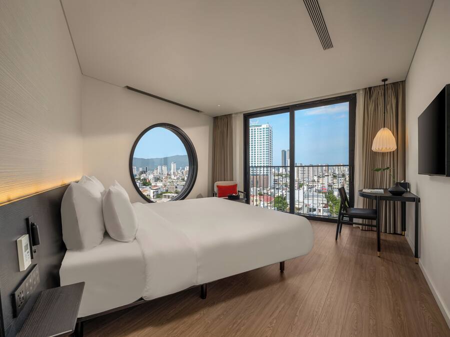 A modern hotel room with a large bed, a desk, a chair, a TV, and a view of the city.