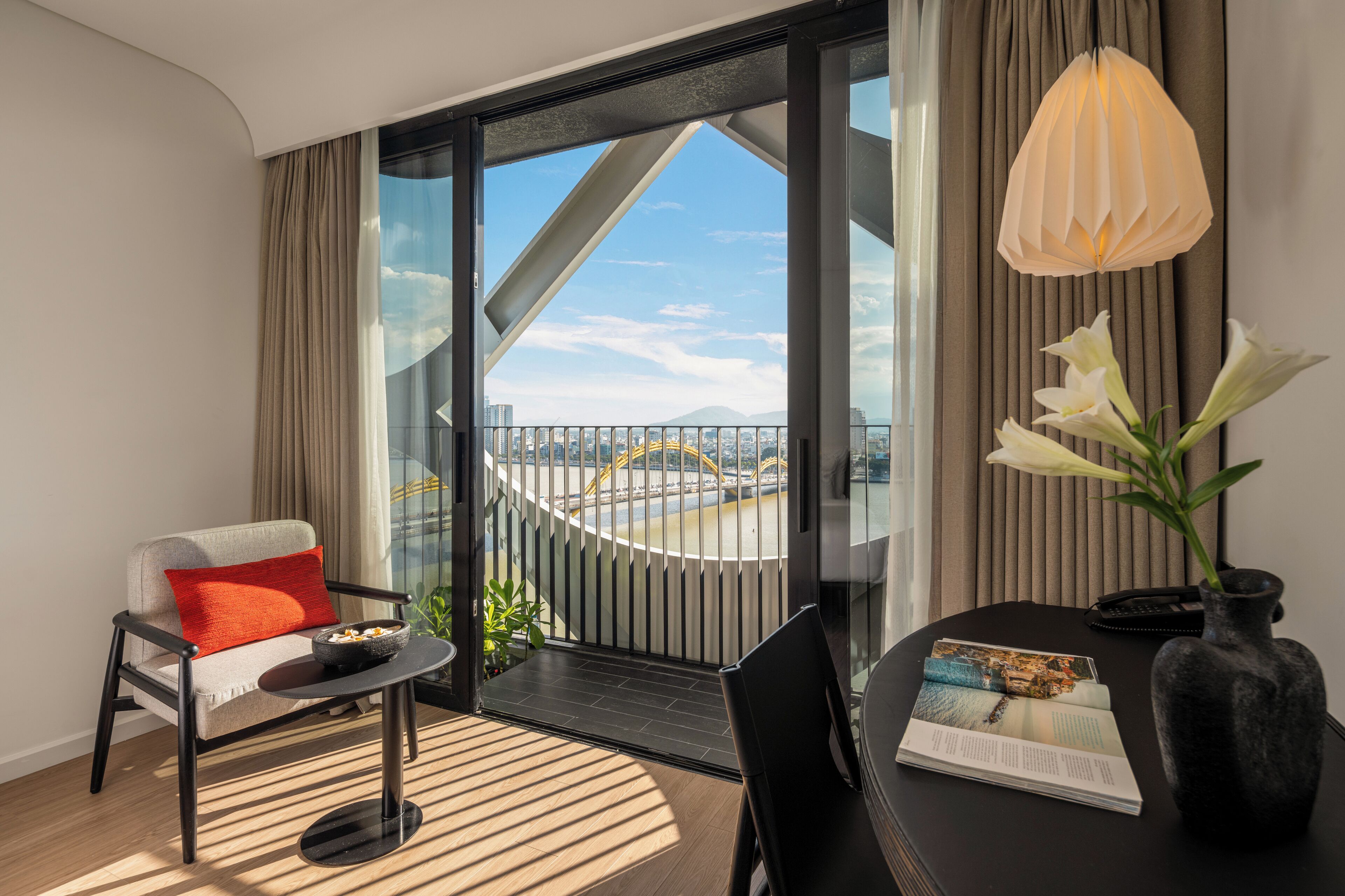 A modern hotel room with a balcony, a sofa, a small table, a vase with flowers, and a magazine on the table.