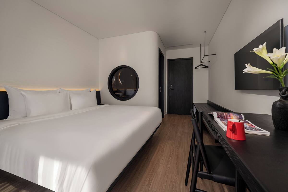 Une chambre d’hôtel moderne avec un grand lit, un bureau sur lequel se trouvent un magazine et une tasse rouge, un miroir rond et un vase rempli de fleurs.