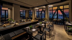 A modern restaurant with a long counter, multiple tables, and large windows offering a city view at dusk. - Caro Premium Danang Hotel (Da Nang)