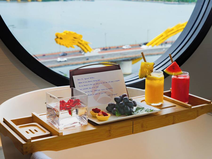A table with a menu, fruit, and drinks, with a view of a bridge and vehicles through a window.