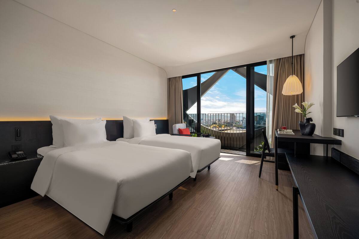 Une chambre d’hôtel moderne équipée de deux lits, d’un bureau, d’une chaise, d’une télévision et d’une grande fenêtre donnant sur la ville.