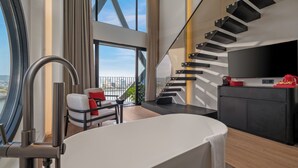 A modern hotel room with a large window, a bathtub, a sofa, and a staircase. - Caro Premium Danang Hotel (Da Nang)