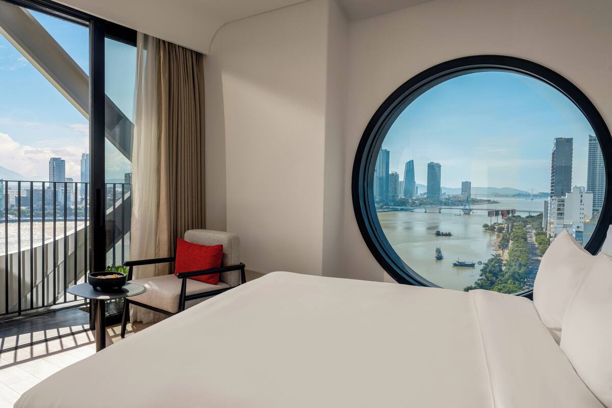 Une chambre d’hôtel avec un grand lit, une fenêtre ronde, une chaise et une table sur laquelle se trouve un saladier rempli de fruits.