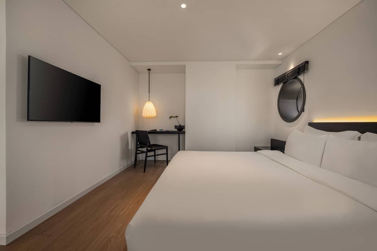 Une chambre d’hôtel moderne dotée d’un grand lit, d’un bureau avec une chaise, d’un téléviseur à écran plat, d’une lampe suspendue et d’un miroir rond.