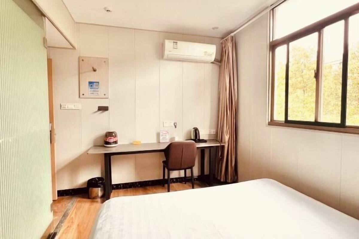 A hotel room with a bed, a desk, a chair, an air conditioning unit, and a window with curtains.