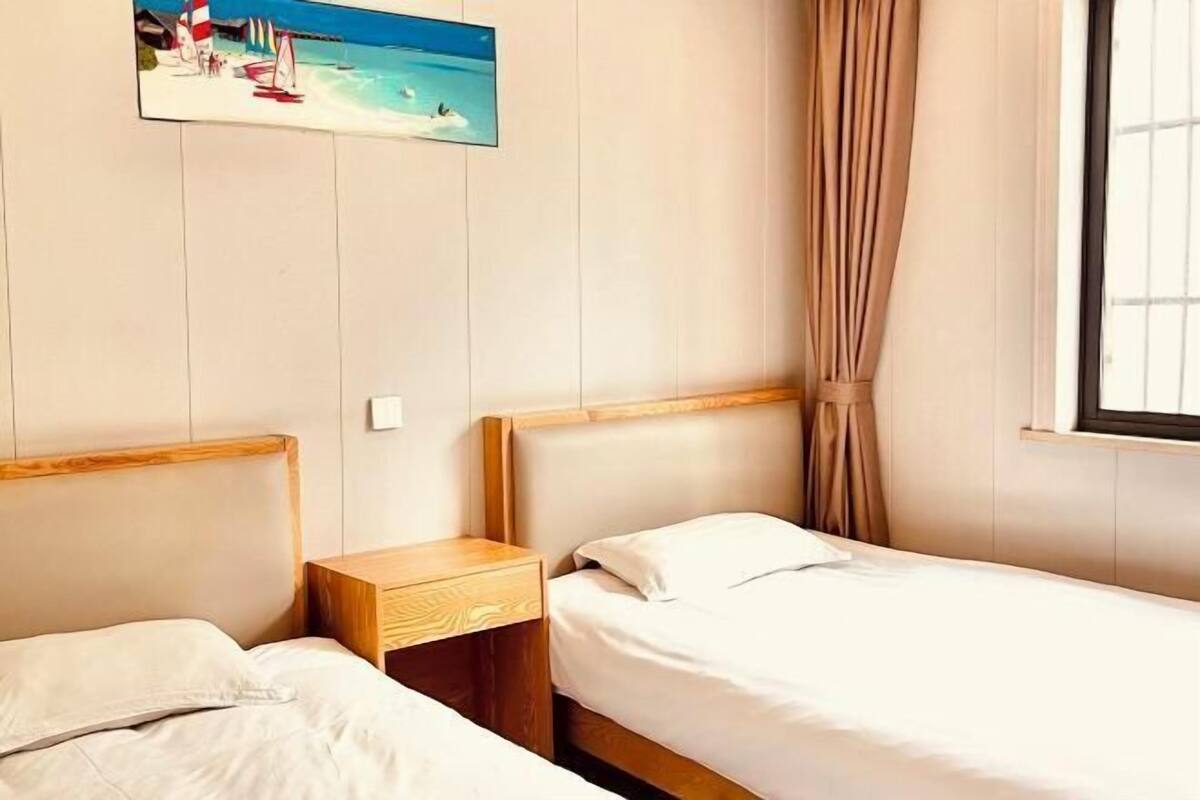 A hotel room with two single beds, a wooden nightstand, a painting of a beach scene, and a window with curtains.