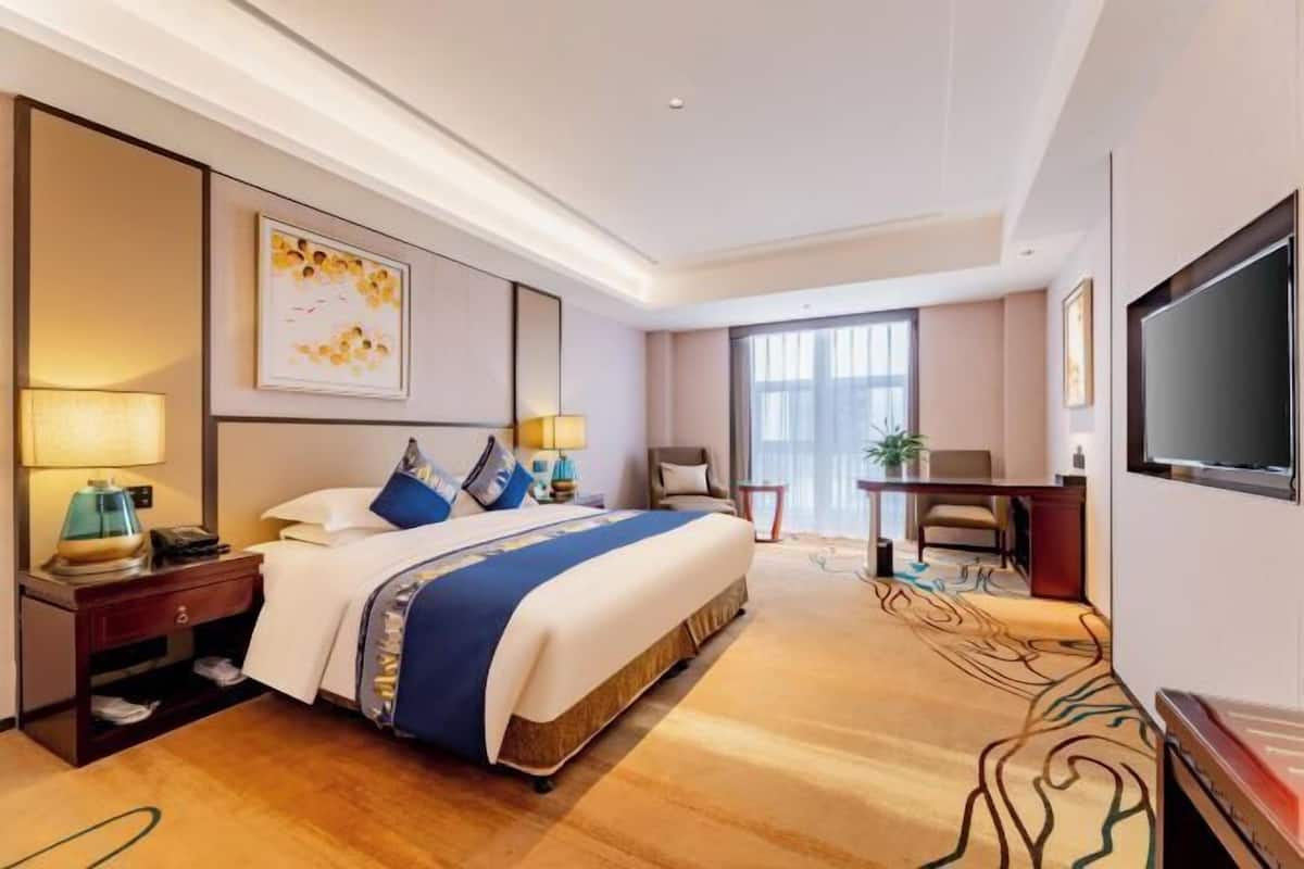Quarto de hotel com uma cama grande, mesas de cabeceira, escrivaninha, uma cadeira, televisão e janela.