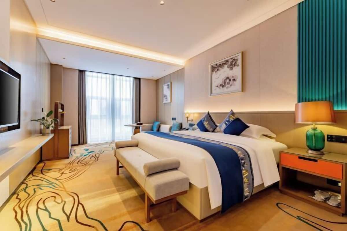 Quarto de hotel moderno com uma cama grande, um banquinho, uma televisão, uma escrivaninha e uma janela com cortinas.