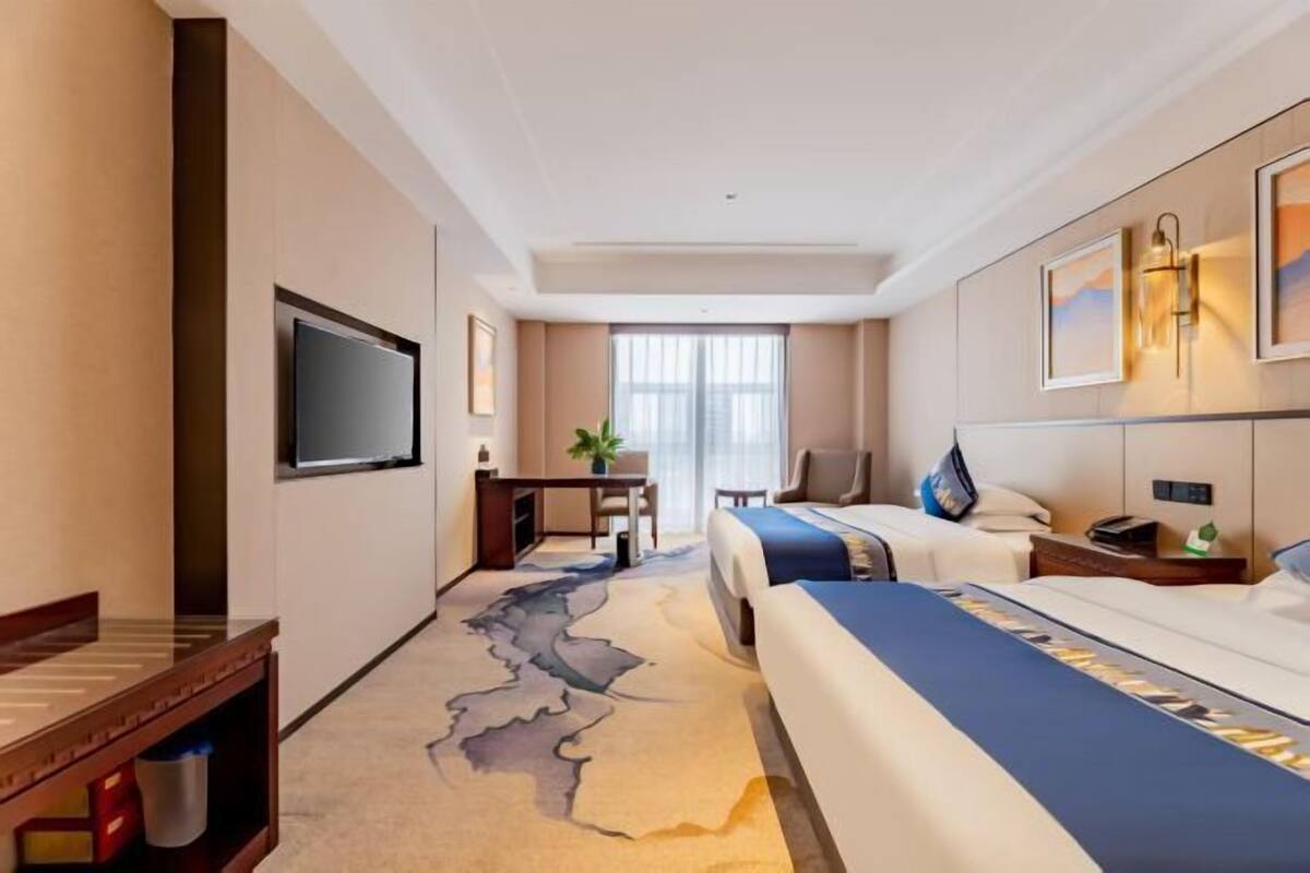 Quarto de hotel com cama, escrivaninha, televisão e uma grande janela.