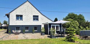 Ein zweistöckiges Haus mit Terrasse, Sitzgelegenheiten im Freien und einem kleinen Garten.