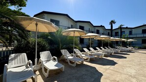 A poolside area with lounge chairs and umbrellas, a building with a flat roof, and palm trees in the background.
