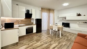 A modern kitchen with white cabinets, a black refrigerator, and a wooden floor. There is a dining area with a table and chairs, and a TV on a white cabinet. - Family Apartments Villaggio Lido B3 - Carraro Immobiliare Jesolo (Cavallino-Treporti)